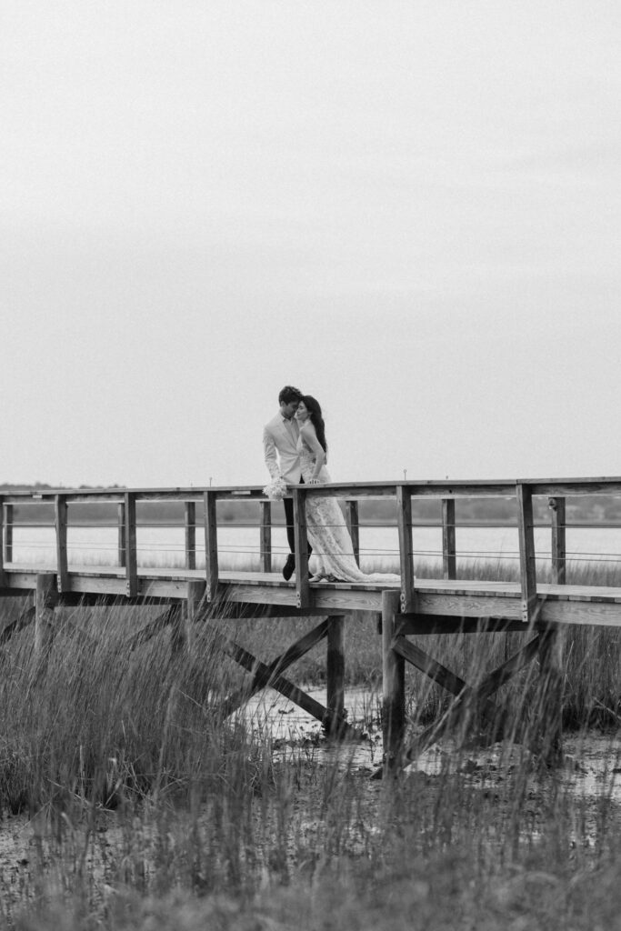 Black-and-white photograph of a couple embracing on a wooden boardwalk surrounded by marshland, offering a serene setting for Charleston SC engagement photos.