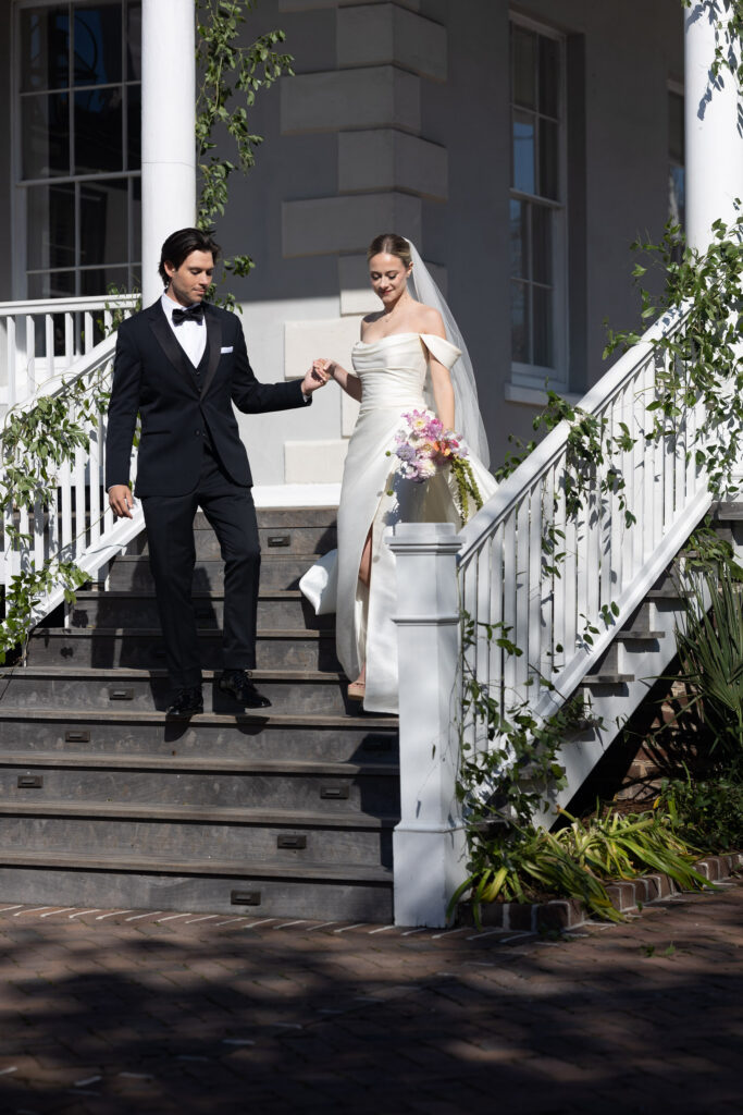 A groom guides his bride down an outdoor staircase wrapped in greenery, photographed at a refined Columbia SC wedding venue.