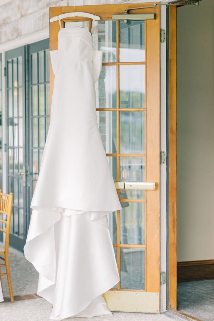 Dunes West Wedding | Stee Garman Photography Fine-art Wedding Photographer | Elegant white strapless wedding dress on a hanger, displayed on a wooden door, with soft natural light creating a serene and romantic atmosphere.