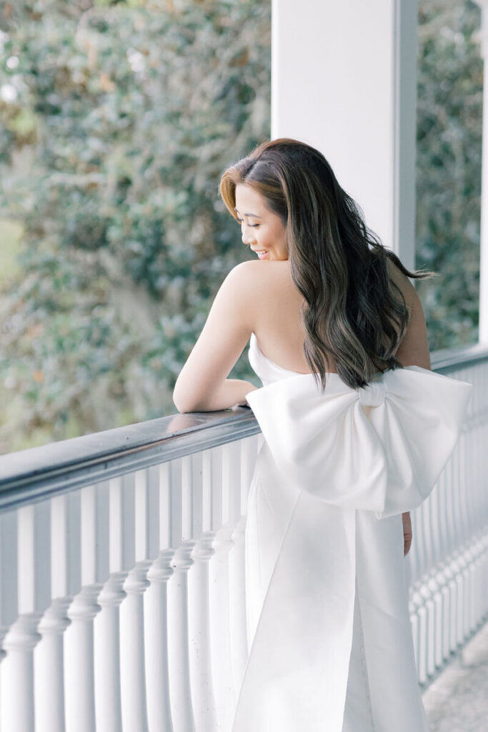 Dunes West Wedding | Stee Garman Photography Fine-art Wedding Photographer | Young woman in a strapless white dress with a large bow at the back stands on a veranda, leaning on the railing, surrounded by lush greenery, smiling softly.