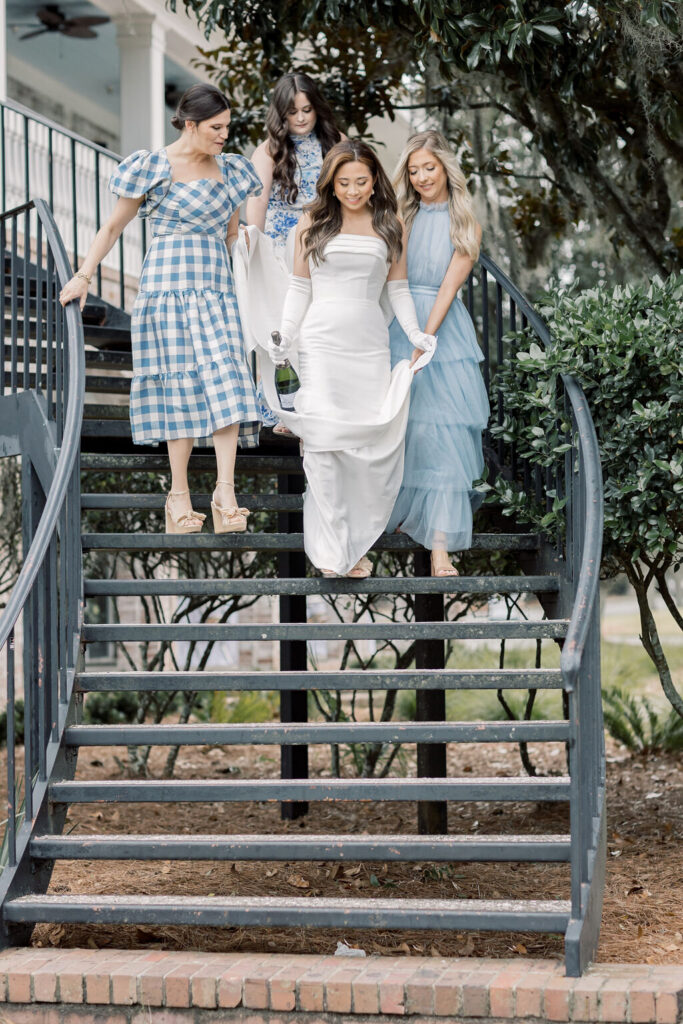 Dunes West Wedding | Stee Garman Photography Fine-art Wedding Photographer | Four women descend a curved staircase outdoors. The bride in a white dress is flanked by bridesmaids in blue dresses, conveying a joyful, elegant mood.