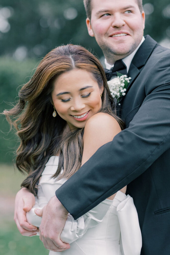 Dunes West Wedding | Stee Garman Photography Fine-art Wedding Photographer | A couple embraces in a tender moment outdoors. The woman in a white dress smiles while the man in a black suit looks away contentedly. Romantic ambiance.