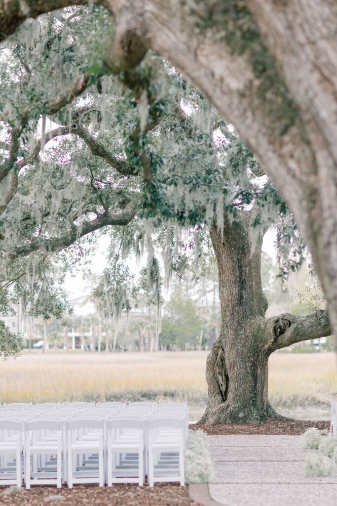 Dunes West Wedding | Stee Garman Photography Fine-art Wedding Photographer | A serene outdoor wedding setup features rows of white chairs under a large oak with hanging Spanish moss. The setting is calm and picturesque.