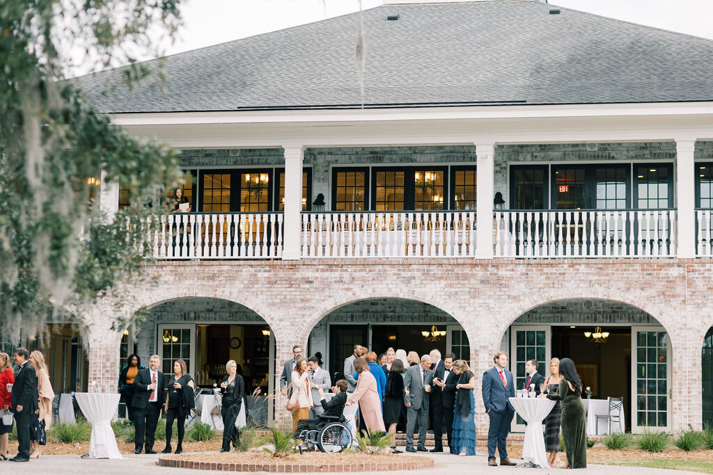 Dunes West Wedding | Stee Garman Photography Fine-art Wedding Photographer | A group of people in formal attire gather outside a large brick building with a balcony. The mood is lively and social, with conversations and mingling.