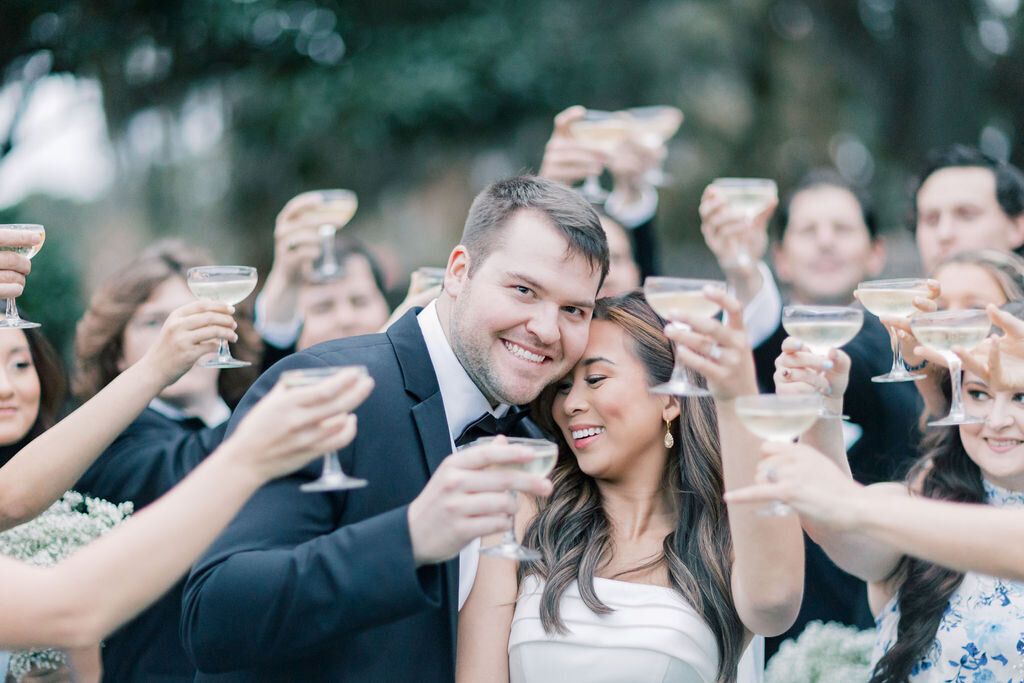 Dunes West Wedding | Stee Garman Photography Fine-art Wedding Photographer | Smiling couple in formal attire surrounded by friends raising glasses in celebration. The atmosphere is joyful and festive, with an outdoor setting.
