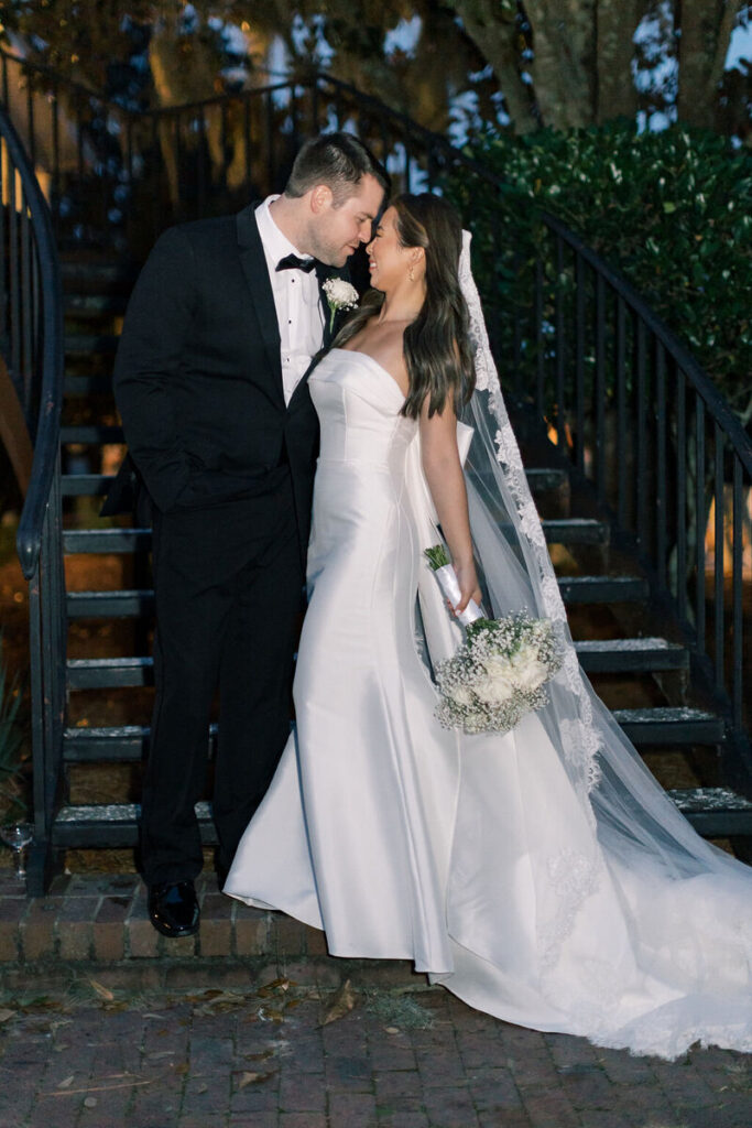Dunes West Wedding | Stee Garman Photography Fine-art Wedding Photographer | Bride and groom share a tender moment on stone steps. Bride in flowing white gown holds bouquet; groom in black tuxedo leans in affectionately, surrounded by evening ambiance.