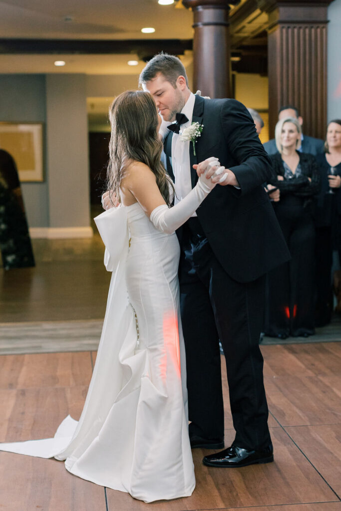 Dunes West Wedding | Stee Garman Photography Fine-art Wedding Photographer | A bride and groom share a tender dance in a warmly lit venue. She wears an elegant white gown with gloves, and he is in a classic black tuxedo. Guests watch fondly.