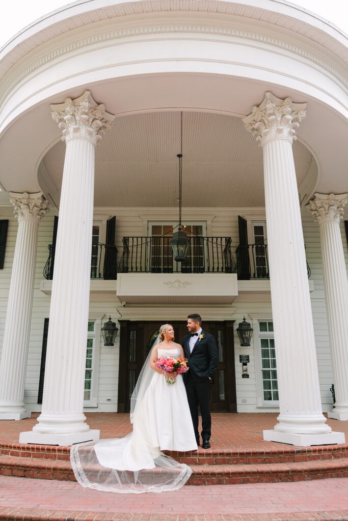 The couple stands beneath the grand white columns at the estate entrance, framed by brick steps and a trailing veil, captured during a timeless Greenville SC wedding.