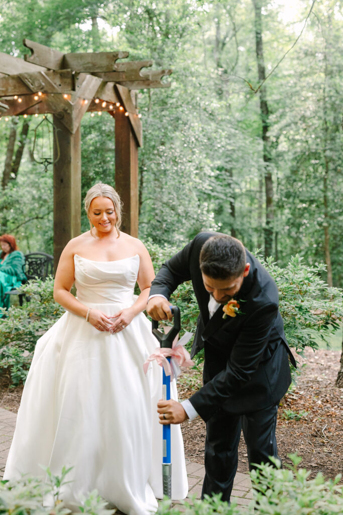 During an outdoor unity ceremony, the groom pours soil as the bride watches beneath a wooden arbor in the trees, telling the story of a heartfelt Greenville SC wedding.