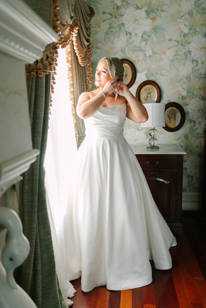 Inside a wallpapered room, the bride fastens her earring near the window in a graceful getting-ready scene by wedding photographers in Charleston SC.