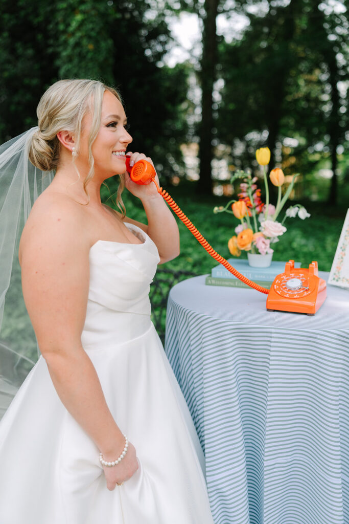 The bride poses beside a bright orange vintage telephone and colorful florals, adding playful personality to this Grand Holland Estate wedding celebration near Greenville SC.