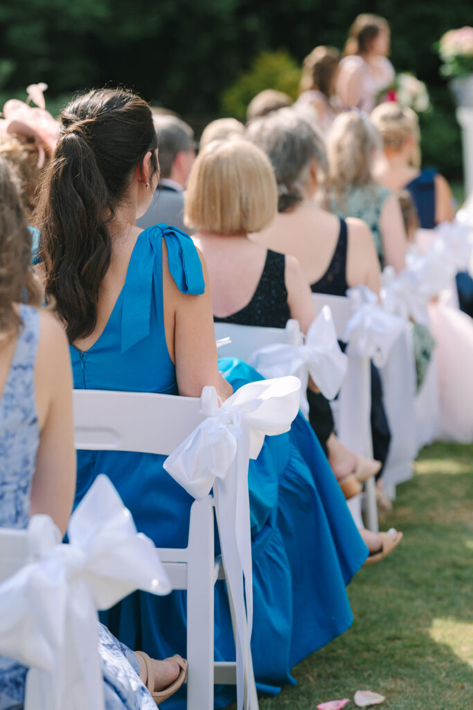 Wedding guests sit along a garden ceremony aisle decorated with white bows as they watch the celebration unfold, a candid moment suited to Charleston wedding photography.