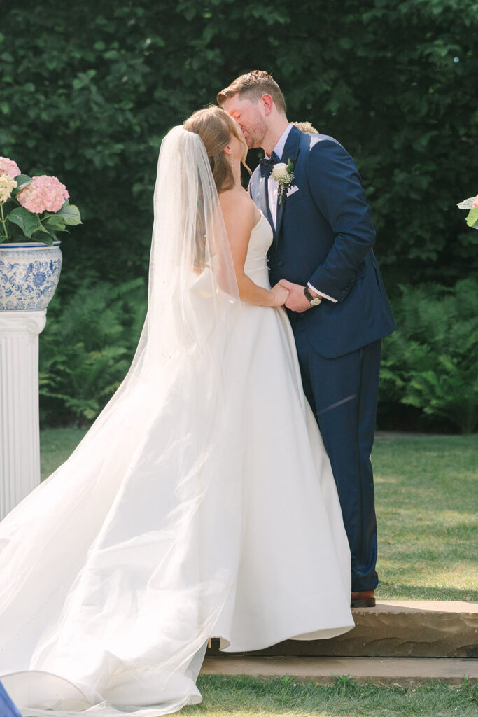The couple shares a quiet kiss beside a garden urn and blooming flowers, framed by greenery during this South Carolina botanical garden wedding.