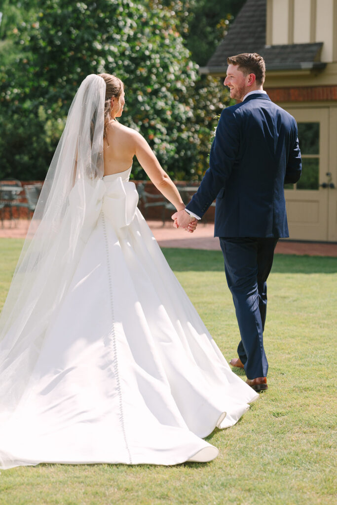 Bride and groom walk hand in hand across a sunlit lawn, surrounded by greenery and the Southern landscape associated with Charleston South Carolina wedding venues.