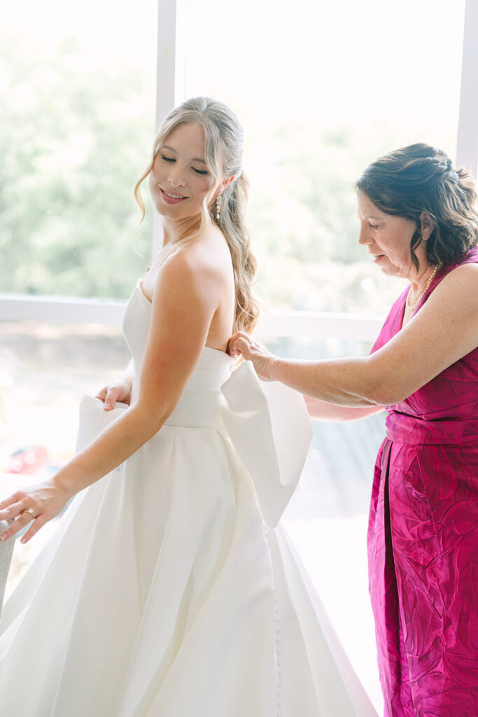 A woman buttons the back of the bride’s gown in bright window light, a tender preparation moment for bridal portraits Charleston SC.