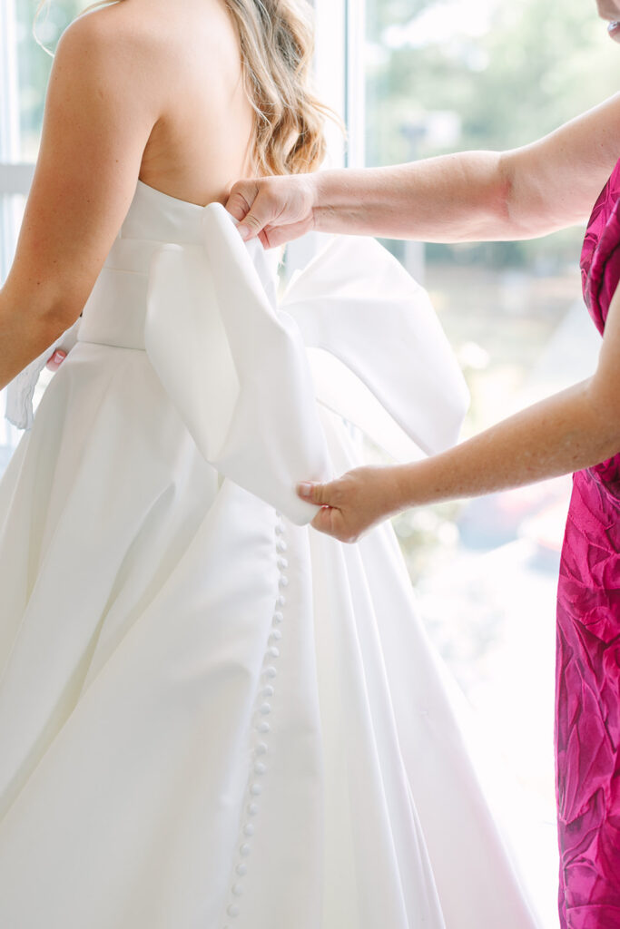 Close-up of a bride’s button-back wedding gown as someone adjusts the fabric beside a bright window, an intimate getting-ready moment at a Charleston wedding venue.