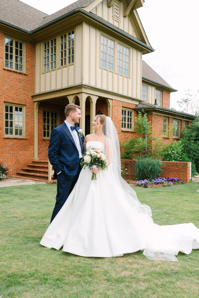 Bride and groom stand together on the lawn outside a brick estate building, her long train spread softly behind them in a timeless portrait from a Botanical Gardens Clemson SC wedding.
