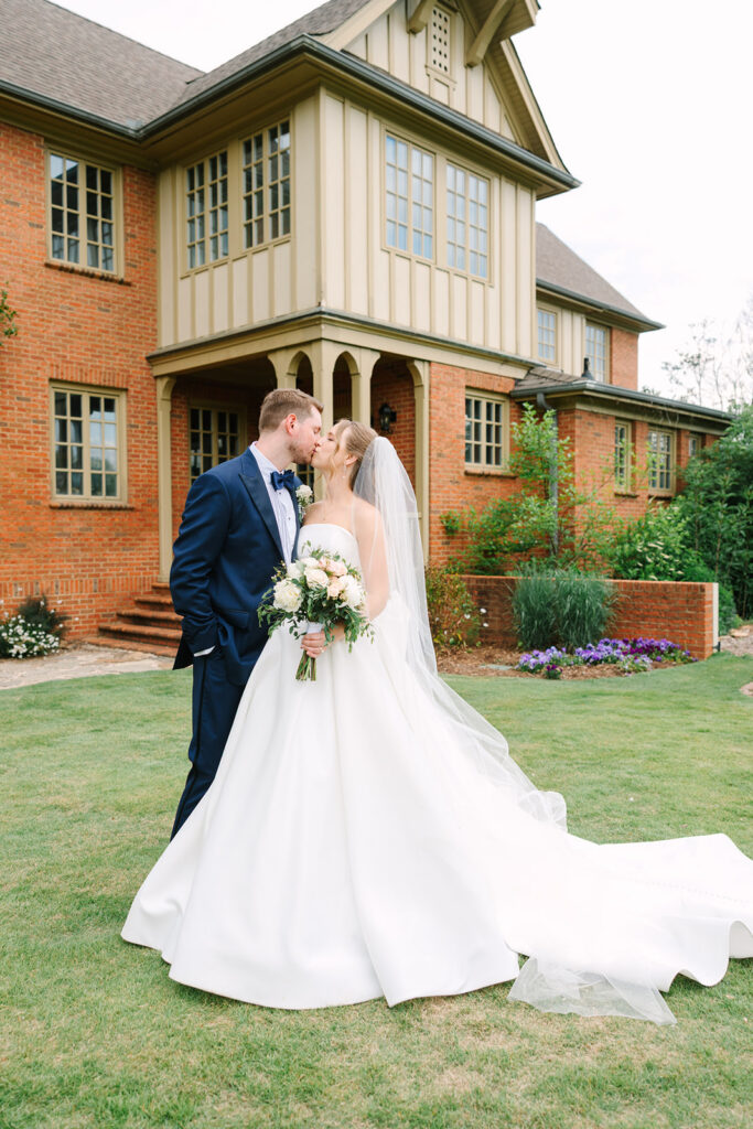 The couple shares a kiss on the lawn in front of a brick-and-stucco home, creating a classic portrait by photographers in Charleston SC.