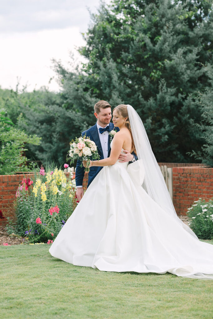 With bright summer flowers and a brick wall behind them, the bride and groom pause for a romantic garden portrait near Clemson, a graceful scene from a garden wedding in Clemson SC.