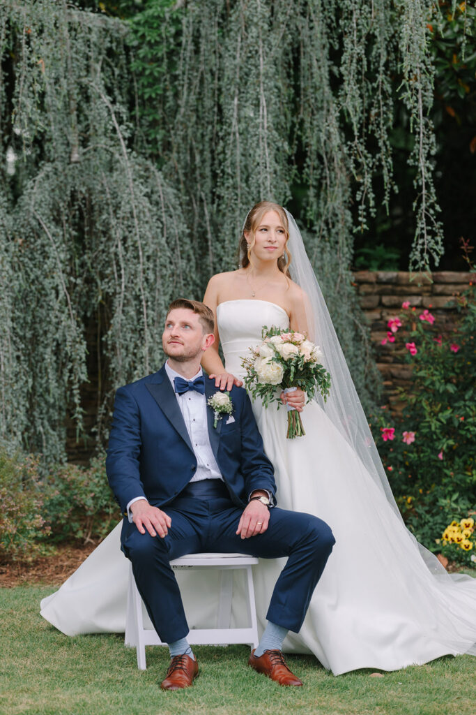 A seated groom and standing bride pose beneath cascading moss and garden greenery at an outdoor wedding venue near Columbia SC.