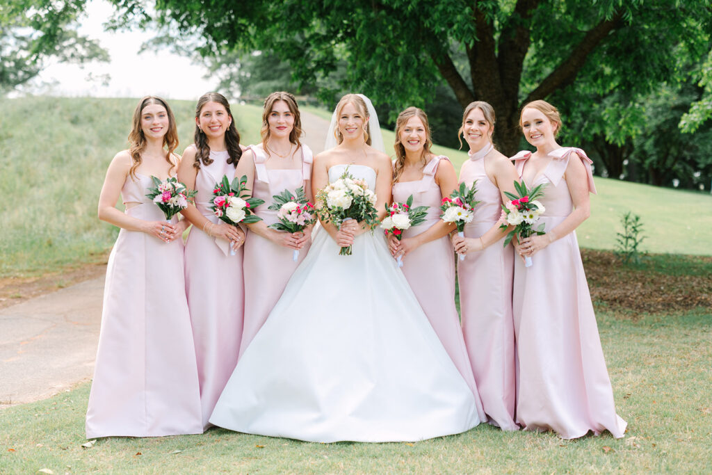 Bride stands beneath a large tree with six bridesmaids in soft blush dresses holding colorful bouquets, a timeless wedding-party portrait suited to Charleston wedding venues with Southern charm.
