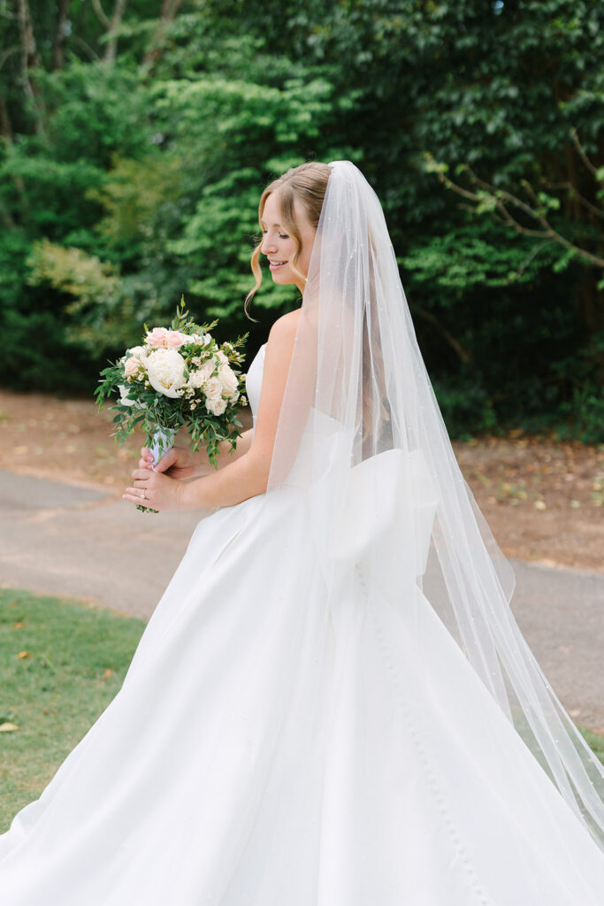 Bride carries a pastel floral bouquet as her long veil and full white gown sweep behind her in a garden setting, a romantic scene at a Charleston wedding venue.