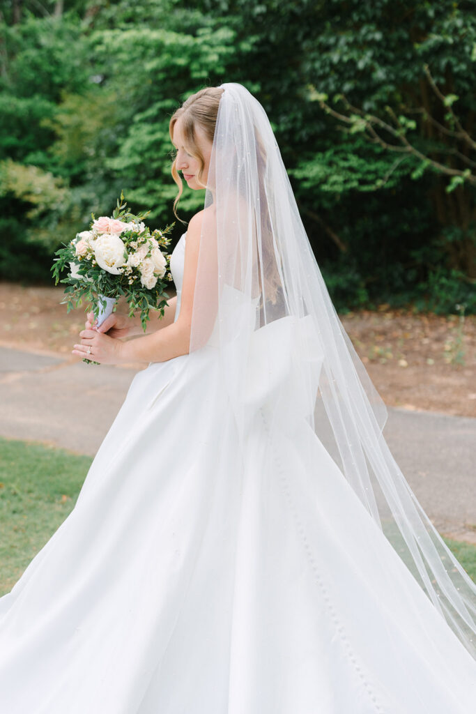 A bride carries a white-and-green bouquet as her full skirt and veil sweep across the lawn, suited to a quiet luxury wedding aesthetic.