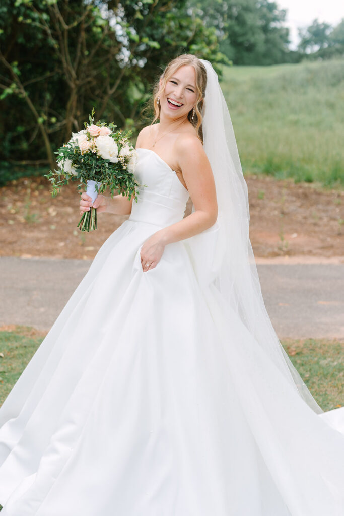 The bride smiles over her shoulder outdoors with bouquet in hand and veil trailing behind her, captured by a Charleston SC wedding photographer.