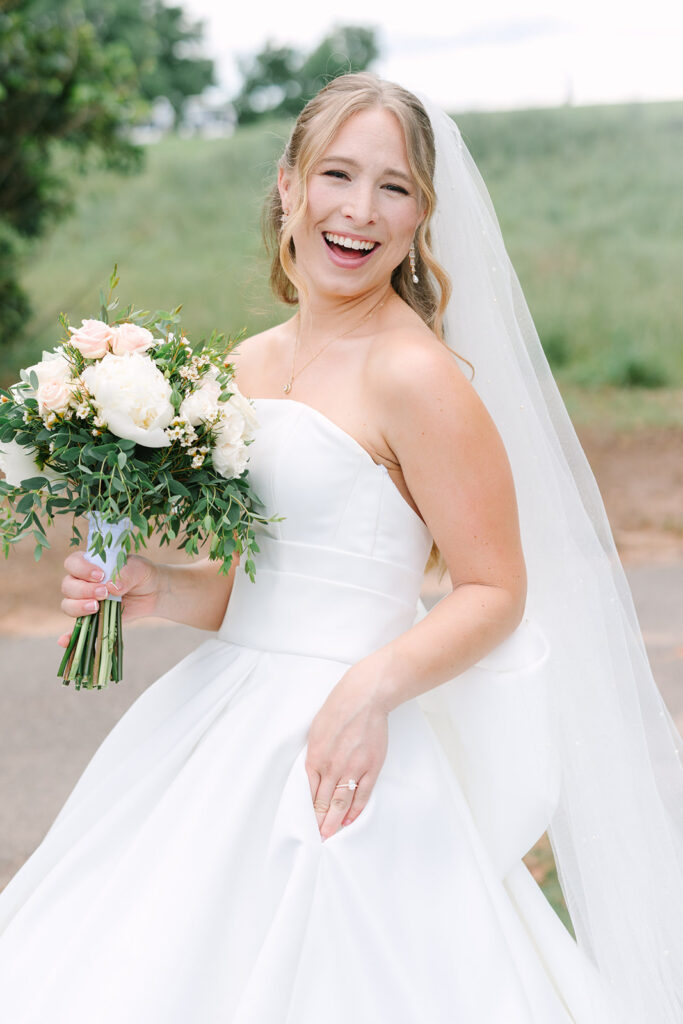 Smiling toward the camera, the bride holds her bouquet in an outdoor portrait with her veil lifting behind her, a lovely moment from a Clemson wedding photographer’s gallery.