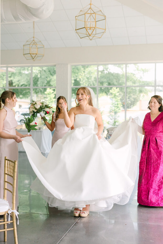 Bridesmaids lift the bride’s gown as she steps across the bright reception space, preserving a playful getting-ready moment from a Botanical Gardens Clemson SC wedding day.