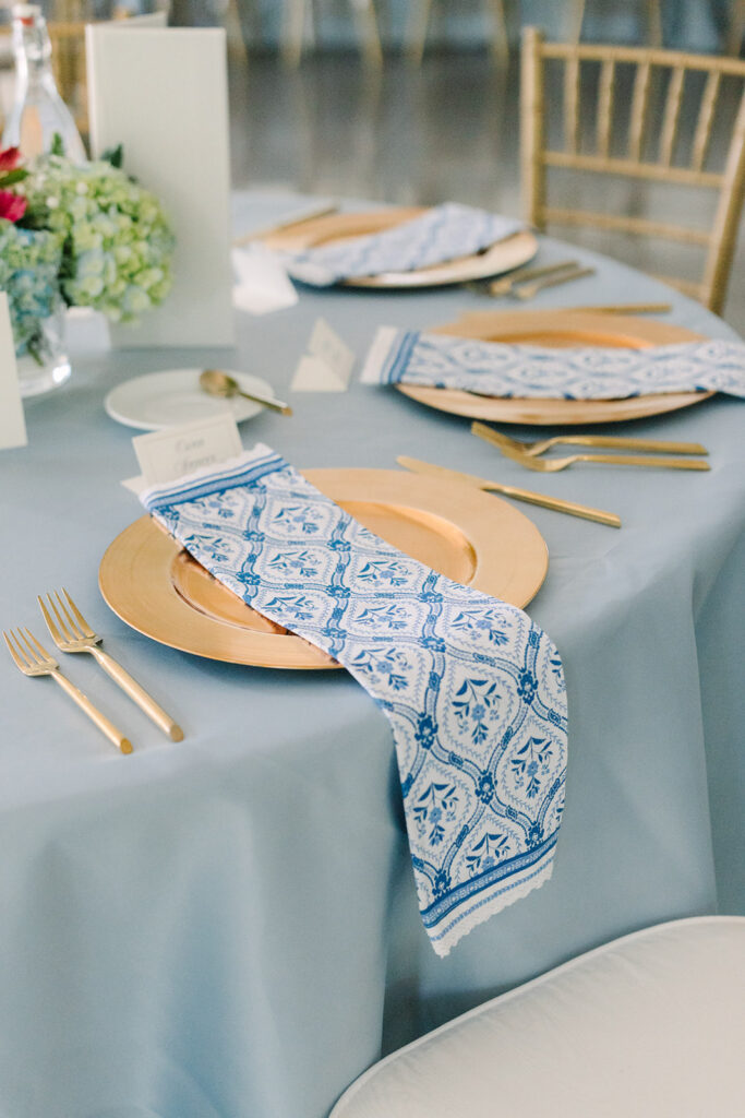 Reception table dressed in soft blue linens with gold place settings and blue-and-white patterned napkins, an elegant detail for a Charleston SC wedding venue.