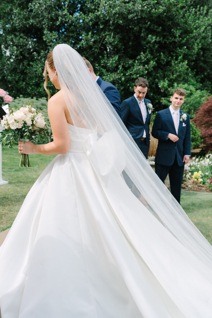 The bride’s veil flows behind her as she turns toward the groom and wedding party on the lawn, a romantic frame from Charleston SC photographers.