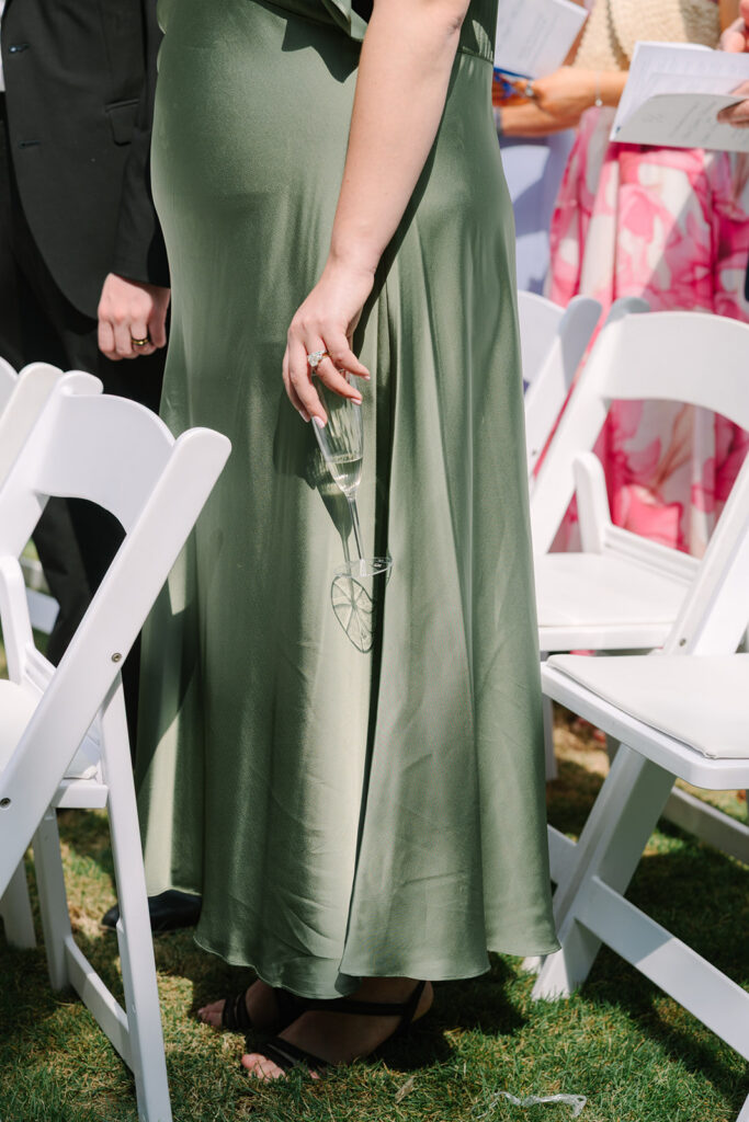 A bridesmaid in a satin green dress walks between white ceremony chairs holding a champagne flute, capturing a candid wedding moment by photographers in Columbia SC.