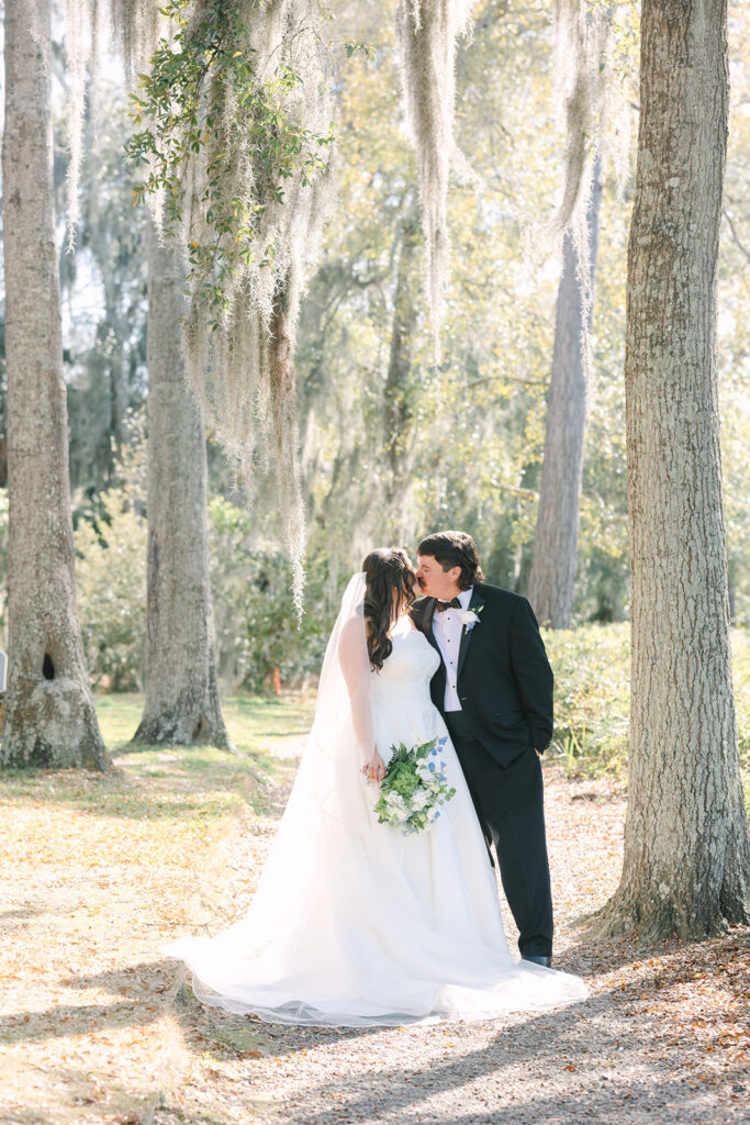 Bride and groom share a quiet kiss beneath tall moss-draped trees, framed by soft afternoon light at Creek Club at Ion.