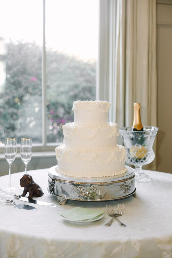 A tiered white wedding cake sits beside champagne and glassware in soft window light, styled for timeless wedding photography in Charleston SC.