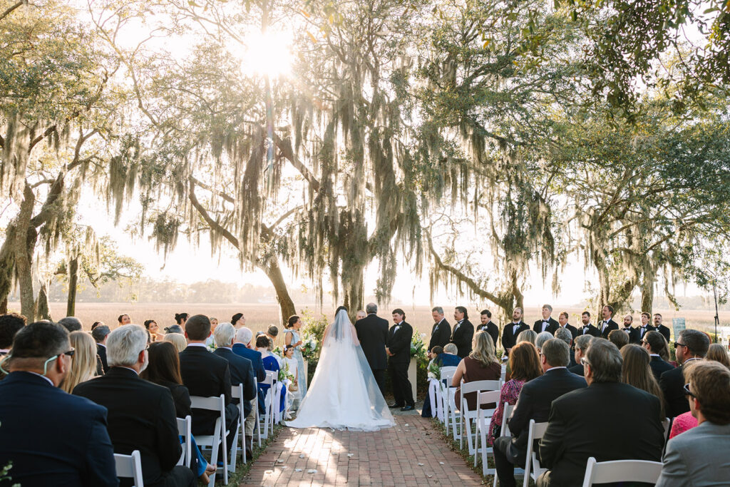 The bride walks toward the ceremony through rows of seated guests while sunlight filters through Spanish moss, a timeless scene from a Creek Club at Ion celebration.