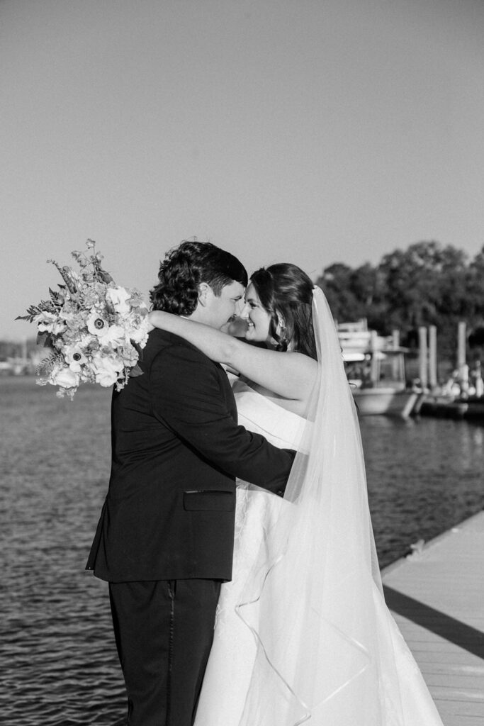 In a black-and-white waterfront portrait, the couple embraces beside the dock with her bouquet lifted over his shoulder, photographed at this Charleston waterfront wedding venue.