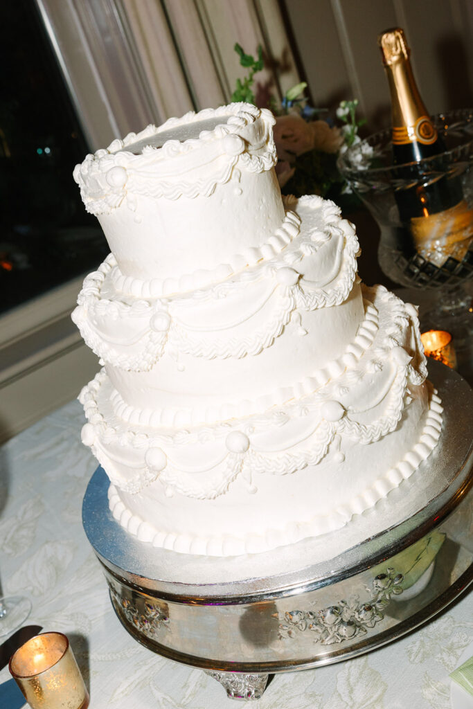 A tiered white wedding cake sits on a silver stand with candlelight and champagne nearby, part of a timeless reception scene from wedding photographers in Columbia SC.
