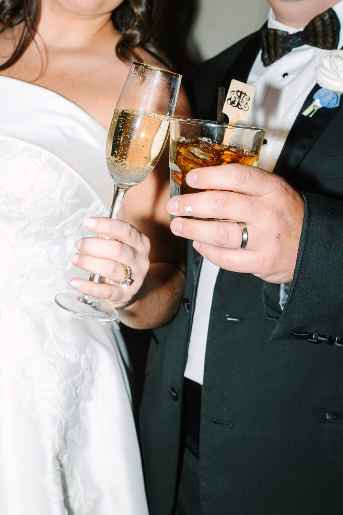 Cropped close, the couple holds champagne and whiskey glasses in a celebratory toast, a refined detail from a Creek Club at I’On wedding day.