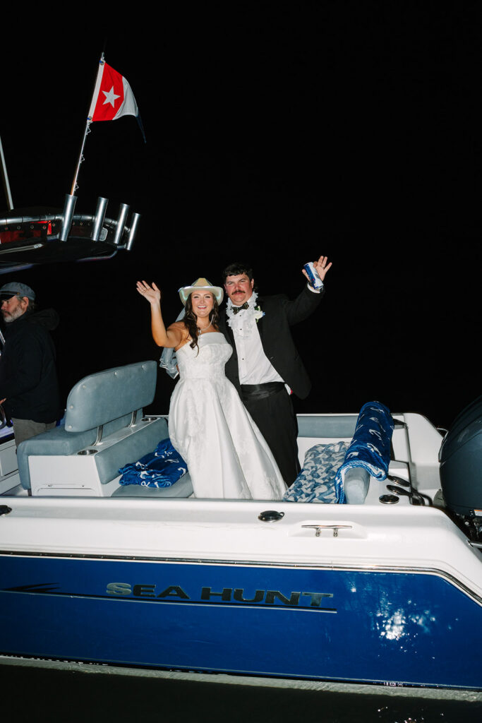 Newlyweds stand on a boat at night with arms raised in celebration beneath a red flag, closing the evening with memorable Creek Club wedding style.