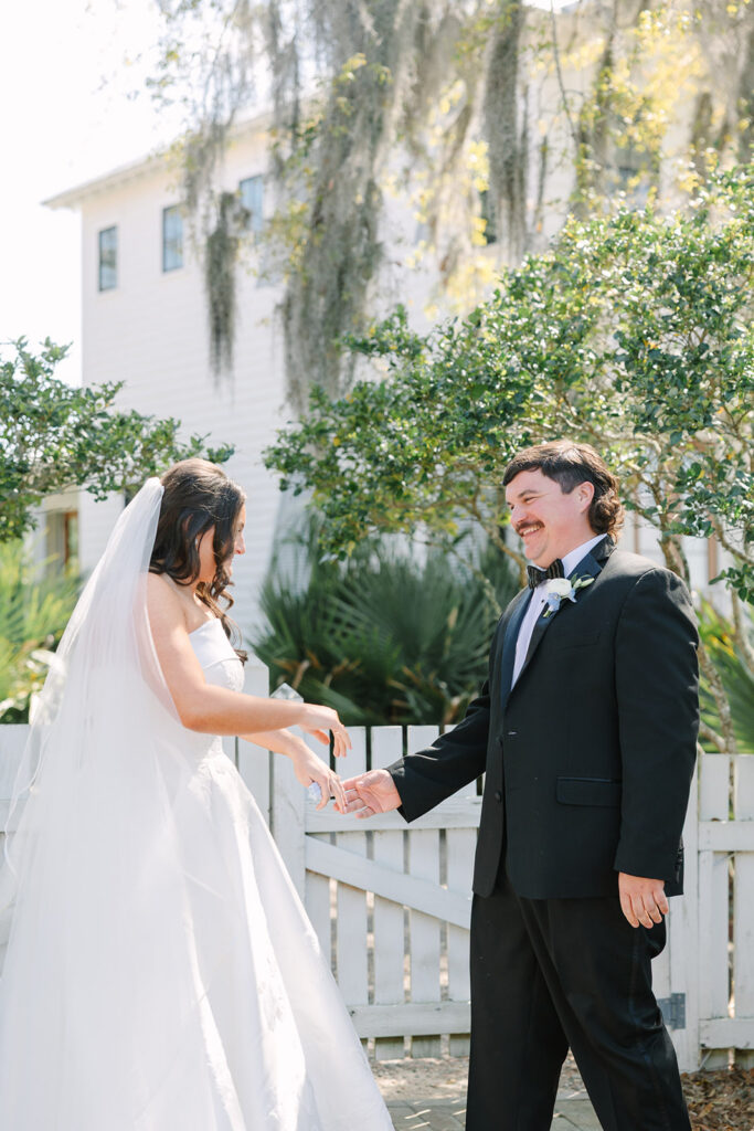 Bride and groom reach for each other across a white garden gate with the historic home behind them, part of a timeless Charleston wedding photographer gallery.